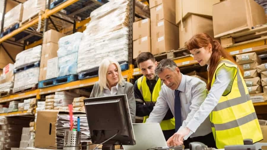 Four staff members, wearing safety vests, collaborate at a desk in a warehouse filled with shelves of boxes and supplies. Two people look at a computer monitor while two others work with papers and a laptop.