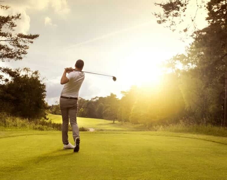 A golfer in mid-swing stands on a green golf course at sunset, with trees and bright sunlight in the background.