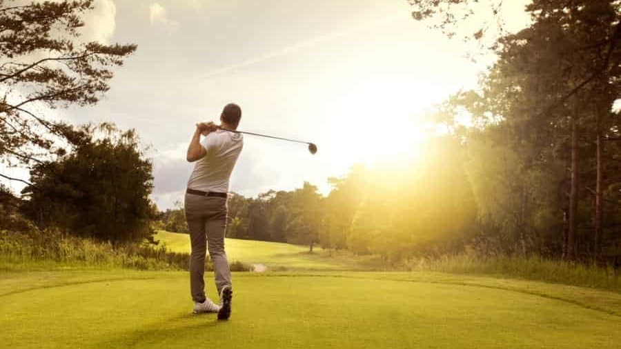 A golfer in mid-swing stands on a green golf course at sunset, with trees and bright sunlight in the background.