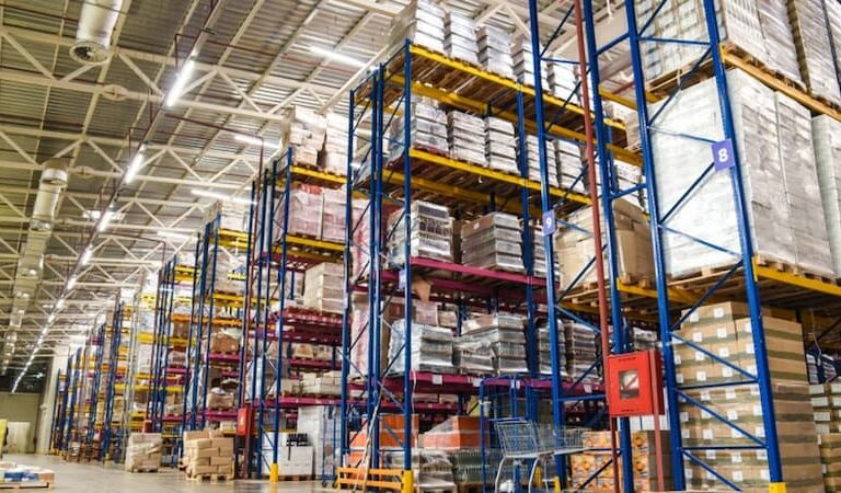 Large warehouse interior with tall blue and yellow racks stacked with pallets and boxes, bright lighting overhead, and organized aisles for storage and distribution.