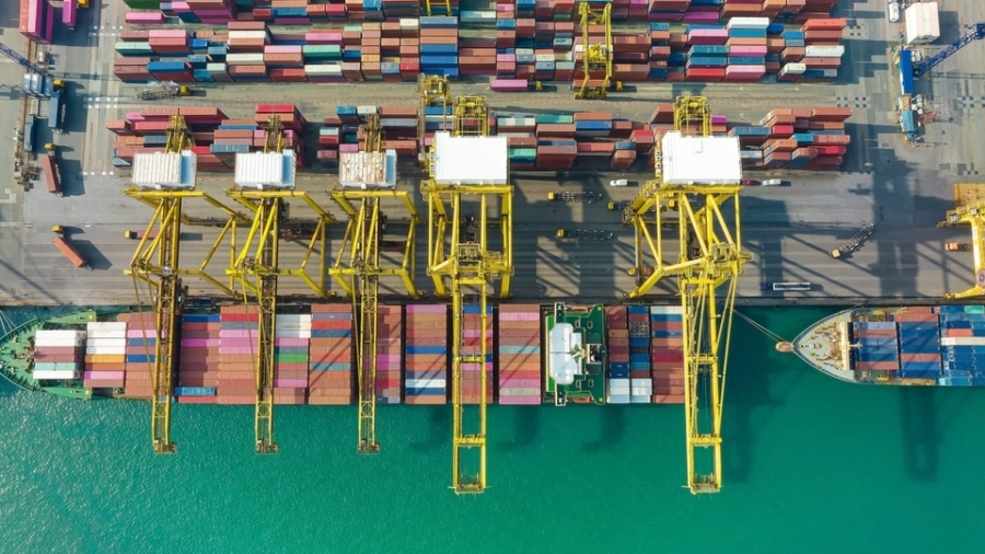 Aerial view of large yellow cranes loading and unloading colorful shipping containers onto cargo ships docked at a busy port, with turquoise water alongside the pier.