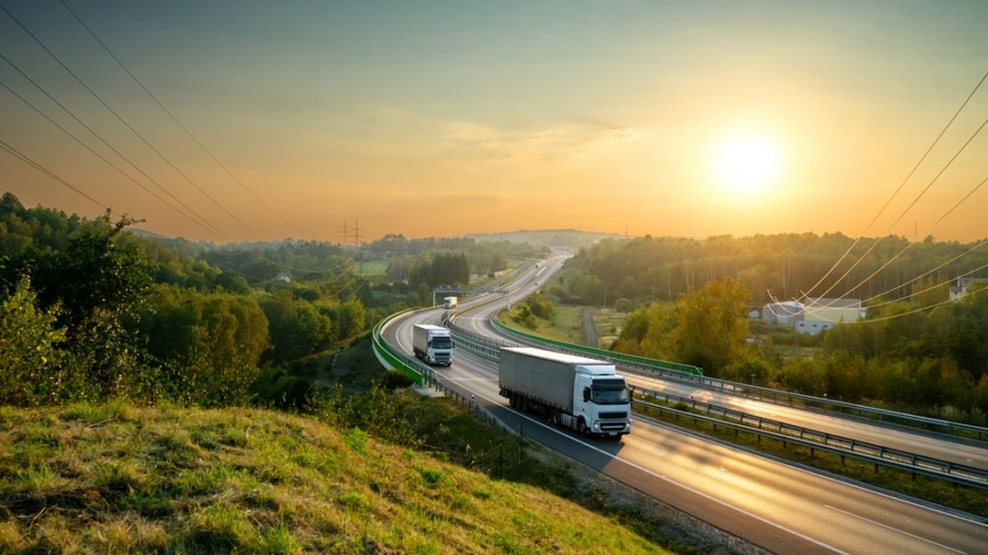 Two semi-trucks drive along a winding highway through green hills at sunrise, the clear sky casting a warm glow—an ideal setting to illustrate a transportation management system business case. Power lines and trees border the serene road.
