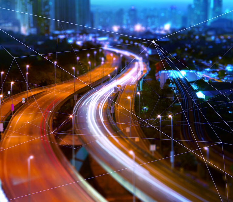 A busy city highway at night with blurred headlights and taillights forming light trails, overlaid with a digital network visualization of glowing lines and nodes, representing smart city technology.