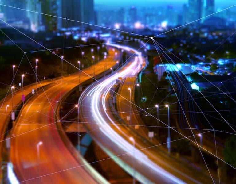 A futuristic cityscape at night with blurred car lights winding through illuminated streets, enhanced by digital network lines overlaying the scene, symbolizing smart city technology and a Transportation Management System driving urban connectivity.