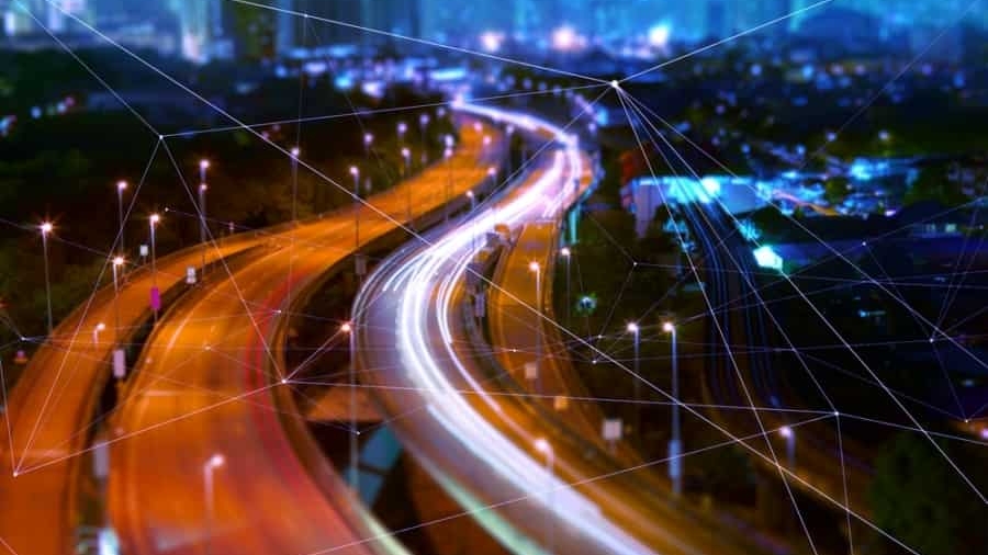 A futuristic cityscape at night with blurred car lights winding through illuminated streets, enhanced by digital network lines overlaying the scene, symbolizing smart city technology and a Transportation Management System driving urban connectivity.