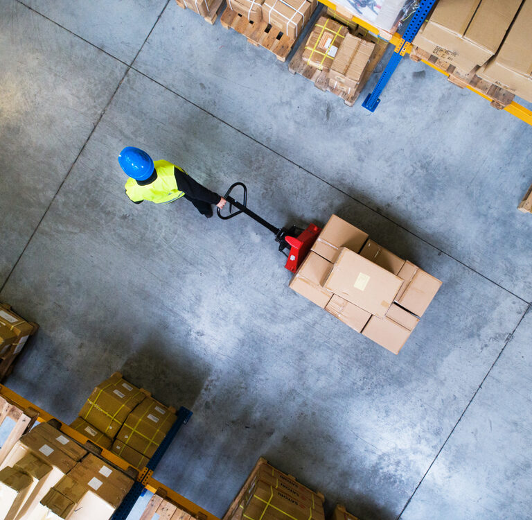 A worker wearing a blue helmet and yellow safety vest moves a pallet of boxes with a pallet jack in a warehouse, surrounded by stacks of cardboard boxes on pallets, viewed from above.