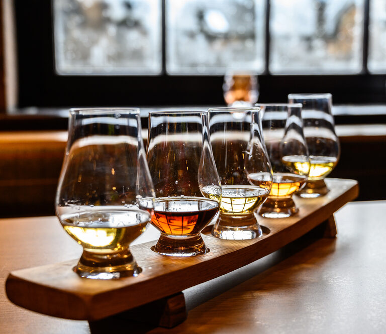 Five glasses of whiskey with varying colors are arranged on a wooden tasting tray, set on a table near a window with blurred outdoor light in the background.