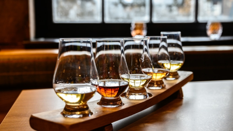 Five glasses of whiskey with varying colors are arranged on a wooden tasting tray, set on a table near a window with blurred outdoor light in the background.