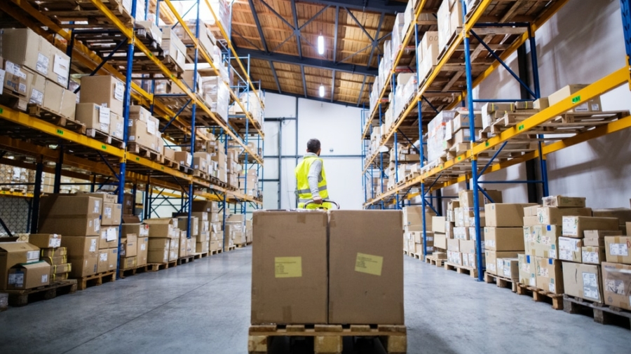A person in a high-visibility vest stands in a warehouse surrounded by tall shelves stacked with cardboard boxes, with two large ab701 boxes on a pallet jack in the foreground.