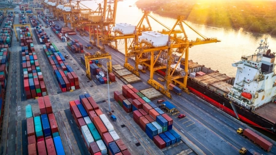 Aerial view of a busy shipping port with large yellow cranes, stacks of colorful cargo containers, and a cargo ship docked alongside a river at sunset—showcasing efficient 3PL operations in action.