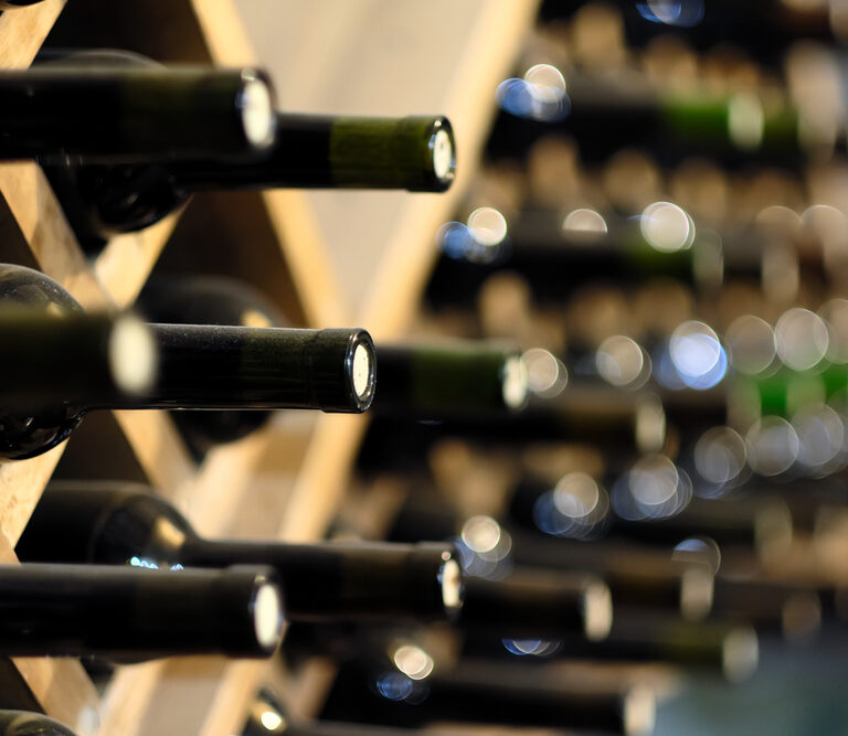 A close-up of wine bottles stored horizontally in a wooden rack, with the bottle necks facing outward. The background shows more wine bottles out of focus.