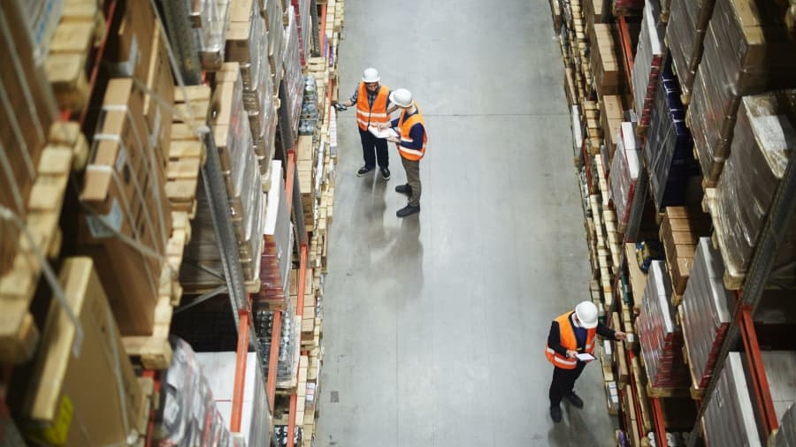 Overhead view of three workers in safety vests and helmets checking shelves and inventory in a warehouse aisle filled with stacked boxes and goods.