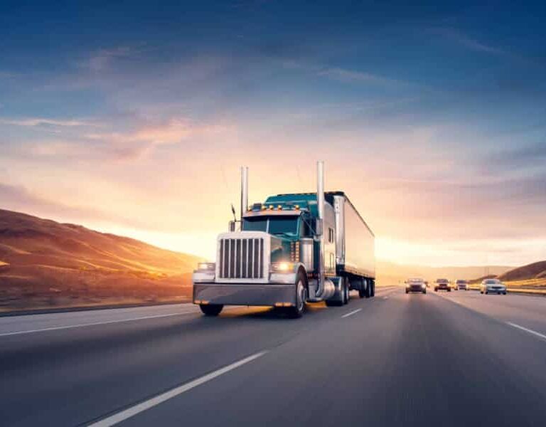 A large semi-truck drives down a multi-lane highway at sunset, with rolling hills in the background and several cars traveling in the opposite direction.
