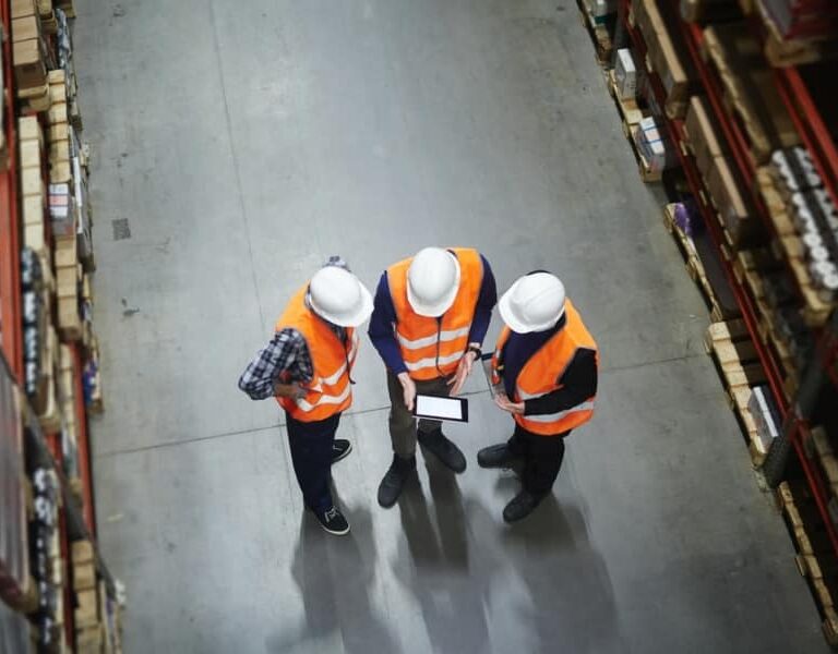 Three people in safety vests and hard hats stand in a warehouse aisle, looking at a tablet together, surrounded by tall shelves filled with boxes and supplies.