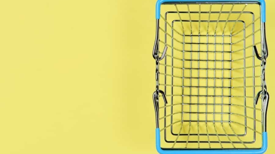 A small, empty metal shopping basket with blue handles is viewed from above against a yellow background, positioned to the right side of the image.