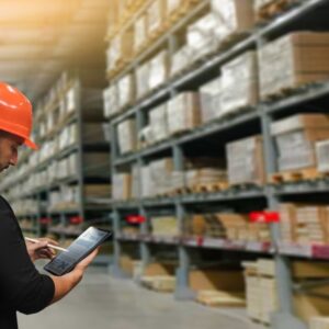 A warehouse worker wearing an orange hard hat and safety vest stands in an aisle, using a tablet, with shelves full of stacked boxes and materials in the background.