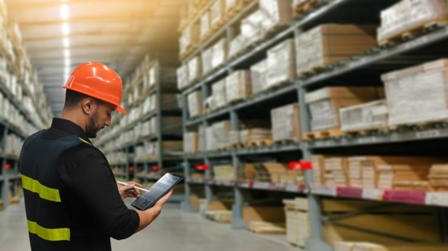 A warehouse worker wearing an orange hard hat and safety vest stands in an aisle, using a tablet, with shelves full of stacked boxes and materials in the background.