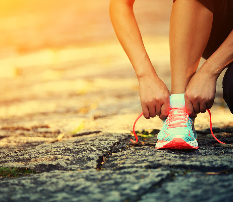 A person kneels on a stone path, tying the bright pink laces of a teal running shoe, with warm sunlight illuminating the scene.