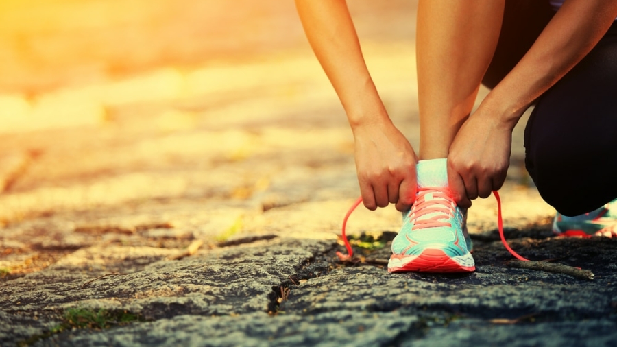 A person kneels on a stone path, tying the bright pink laces of a teal running shoe, with warm sunlight illuminating the scene.
