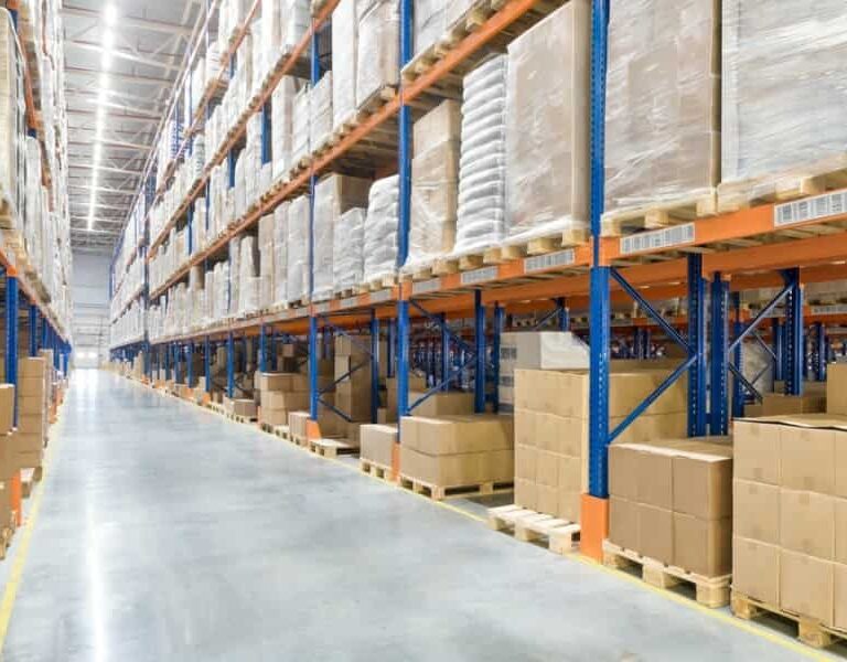 Large warehouse interior with tall shelves filled with stacked boxes and pallets, showcasing organized storage and advanced warehouse automation in a clean, well-lit space along a wide polished concrete aisle.