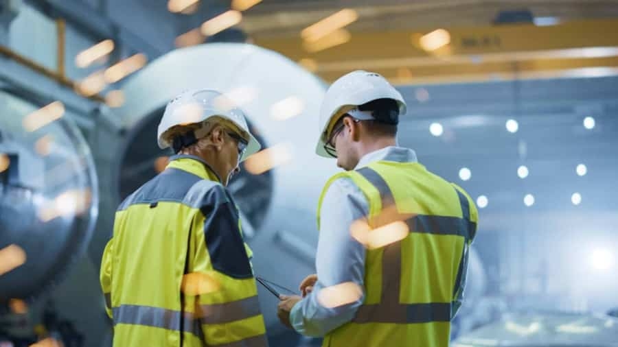 Two workers wearing safety helmets and high-visibility jackets stand in an industrial facility, talking and looking at a clipboard, with bright sparks flying in the foreground.