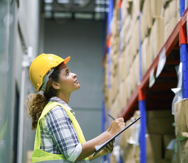 A woman wearing a yellow safety helmet and vest holds a clipboard and pen, looking up at shelves filled with boxes in a warehouse, demonstrating her commitment to warehouse safety.