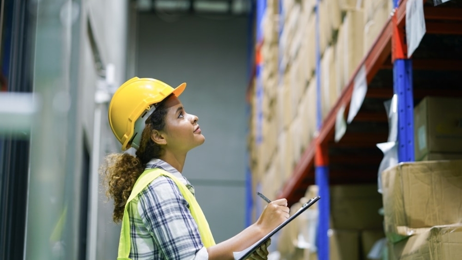 A woman wearing a yellow safety helmet and vest holds a clipboard and pen, looking up at shelves filled with boxes in a warehouse, demonstrating her commitment to warehouse safety.