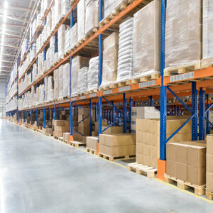 Large warehouse interior with tall blue and orange metal shelving racks stacked with cardboard boxes and wrapped pallets on both sides of a wide, polished concrete aisle. No people are visible.