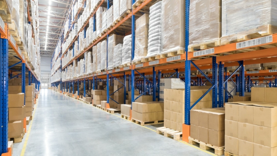 Large warehouse interior with tall blue and orange metal shelving racks stacked with cardboard boxes and wrapped pallets on both sides of a wide, polished concrete aisle. No people are visible.