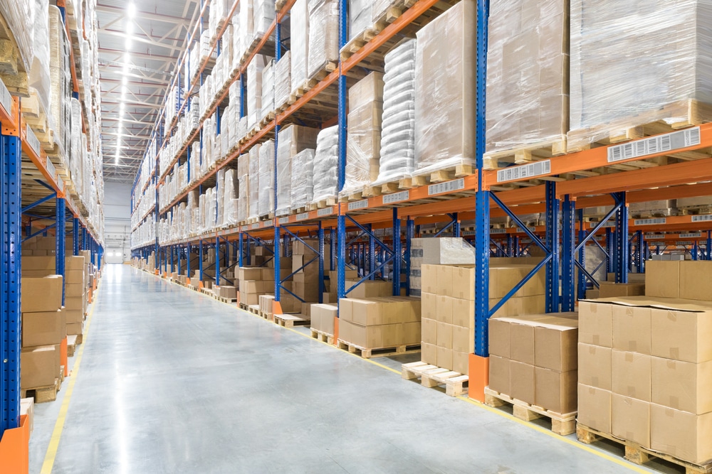 Large warehouse interior with tall blue and orange metal shelving racks stacked with cardboard boxes and wrapped pallets on both sides of a wide, polished concrete aisle. No people are visible.