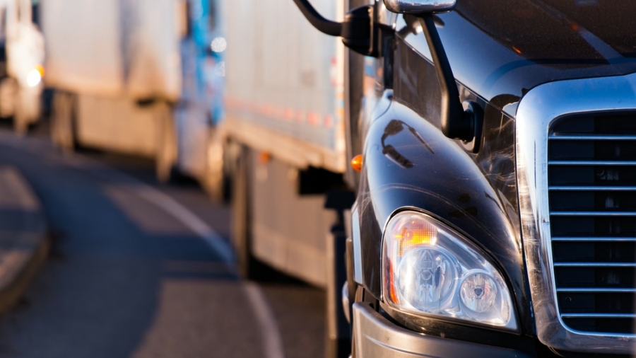 Close-up of the front of a black semi truck on a road, with other trucks lined up behind it, suggesting a convoy or traffic on a highway in the early morning or late afternoon—a vital scene in transportation vulnerable to cyber-attacks.