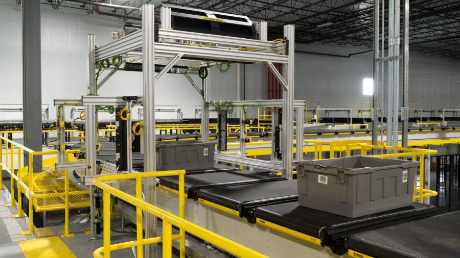 A large industrial warehouse features automated conveyor belts with gray plastic bins and a metal framework, bordered by yellow safety railings. The scene is clean and brightly lit.