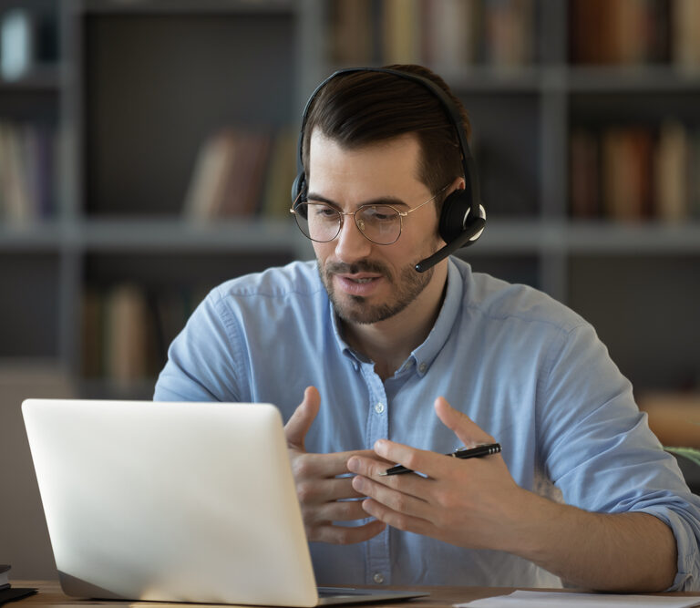 A man wearing glasses and a headset sits at a desk, gesturing as he discusses Microsoft’s Sales Copilot during a video call on his laptop. A bookshelf is visible in the background.