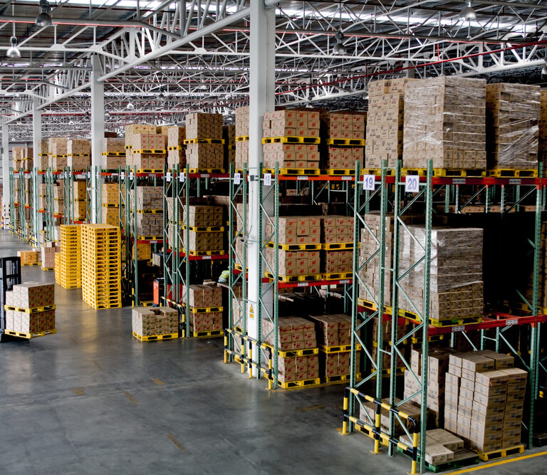 Large warehouse with high shelves stocked with boxes. Two forklifts move through the spacious, well-lit area, transporting goods. Efficient slotting ensures rows of pallets and organized inventory fill the industrial space.