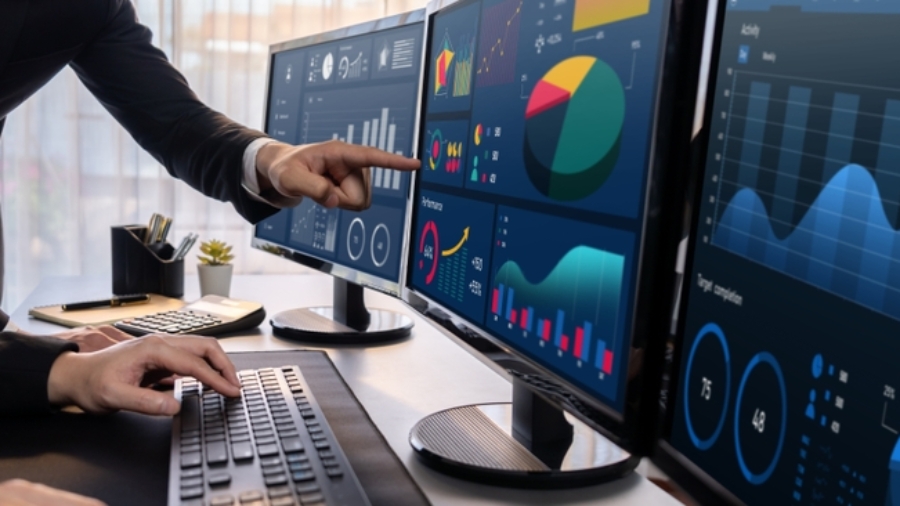 Two people analyze colorful financial graphs and charts powered by Microsoft Business Solutions on three large monitors in a modern office. One types on a keyboard while the other points at the screen, discussing data trends.