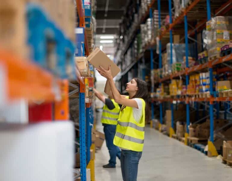 A woman wearing a yellow safety vest reaches for a box on a high shelf in a large warehouse, with other workers and shelves stocked with various products visible in the background.