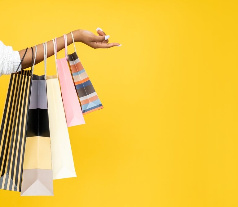 A person’s outstretched arm holds several colorful shopping bags against a bright yellow background. Only the arm and hand are visible, and the bags have various stripes and pastel colors.