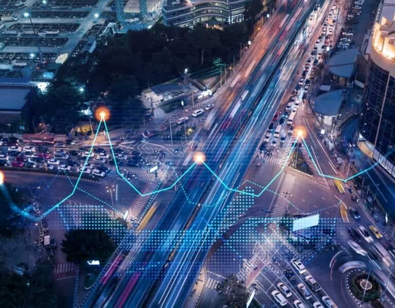 A busy city intersection at night with blurred car lights and overlaid digital graphics, including a glowing line graph and data points, representing traffic analysis or smart city supply chain technology selection.