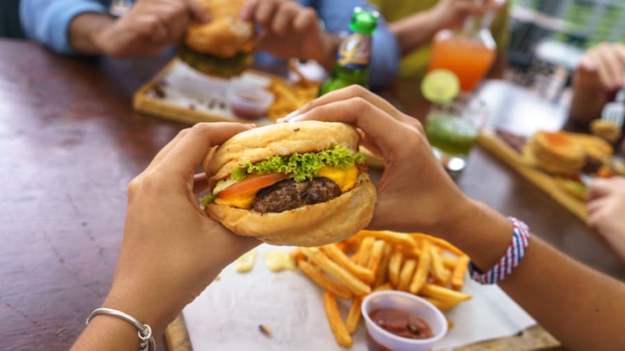 A person holds a burger with lettuce, tomato, and cheese over a tray of fries and ketchup, while others eat burgers and drink beverages at a wooden table.