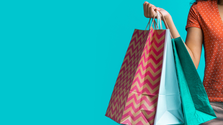 A person wearing an orange polka dot shirt holds several colorful shopping bags against a bright turquoise background. Only the arm and part of the torso are visible.
