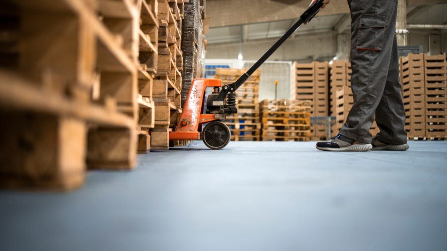 A worker in gray pants and shoes uses a hand pallet jack to move wooden pallets inside a warehouse with stacks of pallets in the background.