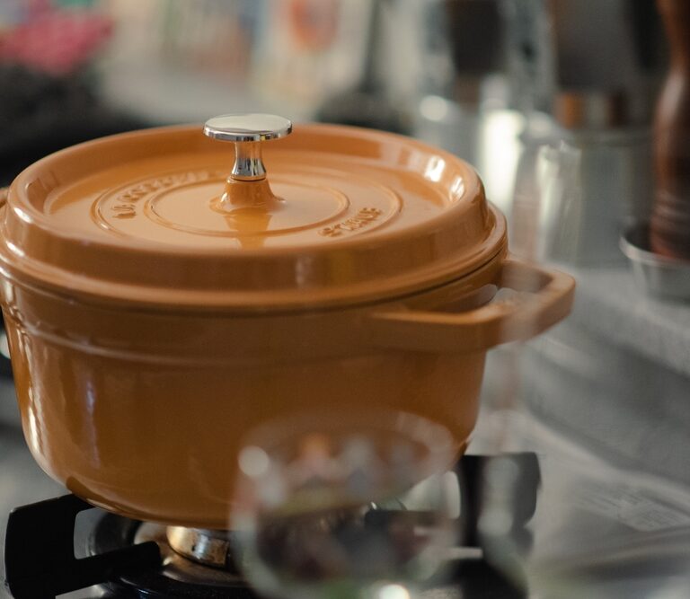An orange enameled cast iron pot with a lid sits on a gas stove burner in a kitchen, with blurred kitchen items and utensils in the background.