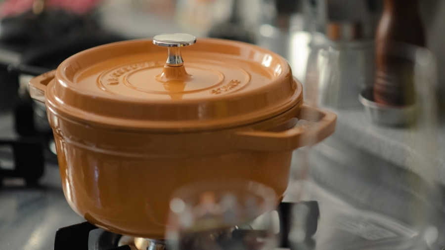 An orange enameled cast iron pot with a lid sits on a gas stove burner in a kitchen, with blurred kitchen items and utensils in the background.