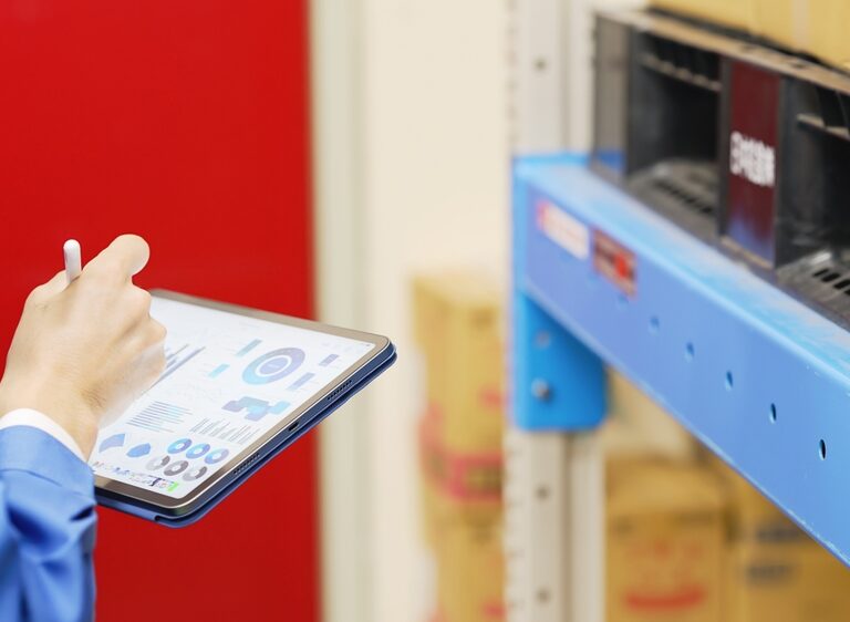 A person in a blue uniform holds a tablet displaying graphs and charts, using a WMS to conduct inventory or inspection in a warehouse filled with shelves and cardboard boxes.