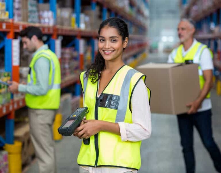 A smiling woman in a yellow safety vest holds a barcode scanner in a warehouse, with shelves of boxes behind her and two workers in vests handling inventory.