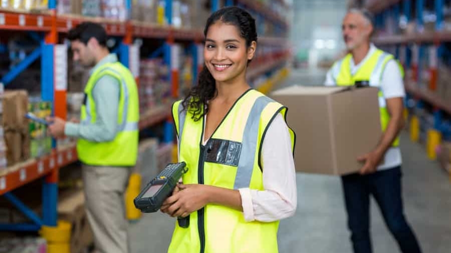 A smiling woman in a yellow safety vest holds a barcode scanner in a warehouse, with shelves of boxes behind her and two workers in vests handling inventory.