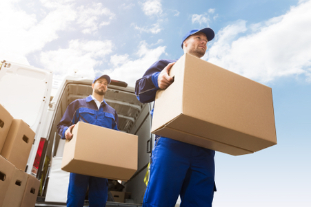 Two workers in blue uniforms carry cardboard boxes from a white van on a sunny day, with a clear blue sky and scattered clouds in the background.