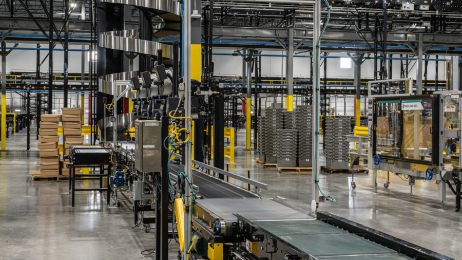 A modern warehouse interior featuring slam solutions, with conveyor belts, stacked cardboard boxes, metal shelves, and various machinery, all under bright industrial lighting.