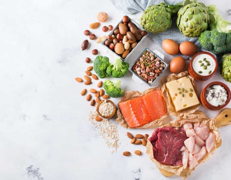 Assorted healthy foods including raw salmon, beef, chicken, cheese, eggs, nuts, seeds, broccoli, artichokes, yogurt, and legumes arranged on a light surface with space on the left.