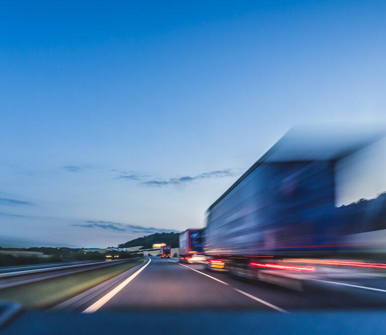 A blurred truck speeds along a highway at dusk, with a clear blue sky and distant hills visible in the background, capturing the essence of efficient transportation sourcing.